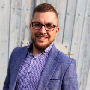 Smiling man in a blue blazer and checkered shirt posing outdoors against a wooden fence (professional headshot).