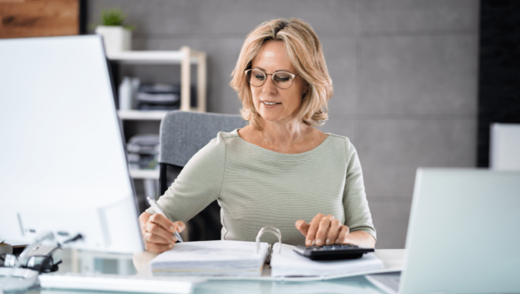 Woman in a light green top sits at a desk in an office, writing in a binder with a pen while using a calculator.