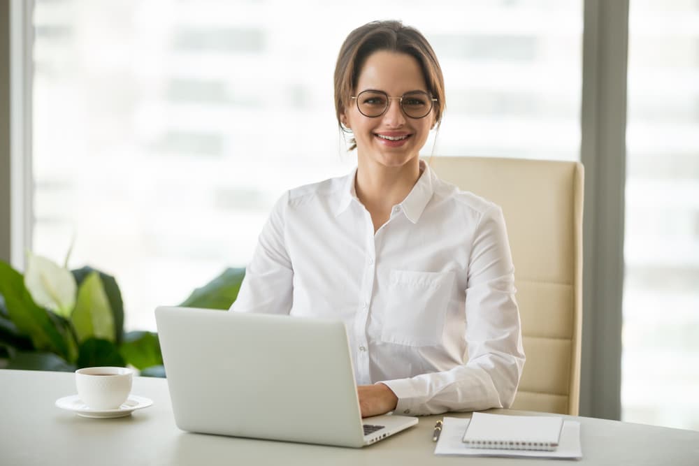 Woman in a white shirt sits at a desk with a laptop, smiling at the camera in a bright office setting.
