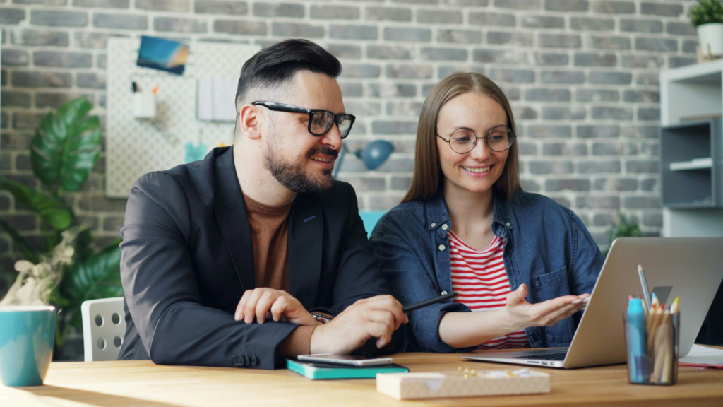 Two colleagues, a man with glasses and a woman with round glasses, sit at a desk discussing over a laptop in a bright office.